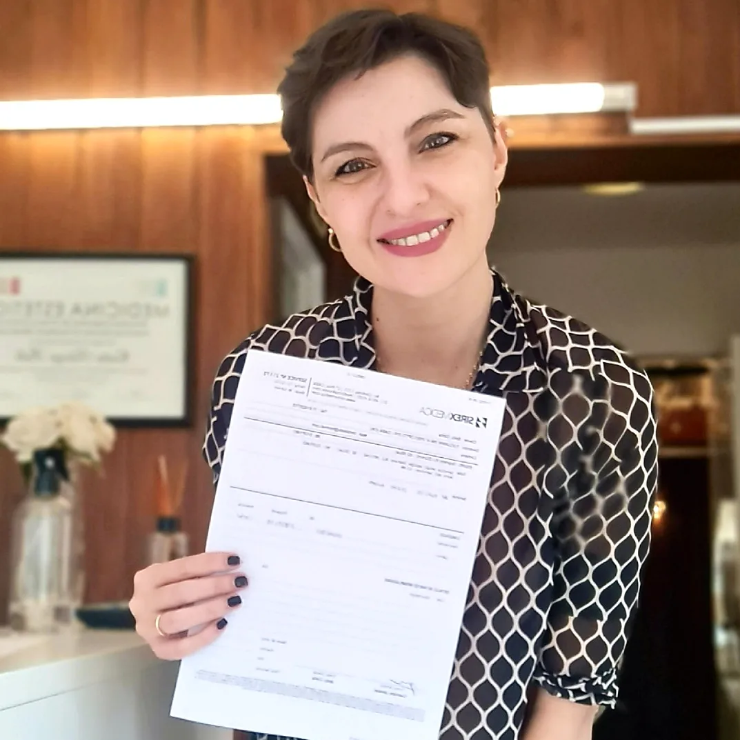 La Dra. Cintia Bello sonriendo en su consultorio de paredes de madera, con flores blancas y su diploma de Medicina Estética detrás